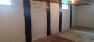 Basement room with white concrete block walls, reinforced by vertical black stripes. Two small windows let in light, creating a stark, industrial feel.