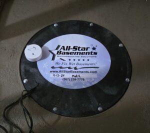 Circular black sump pump cover on a concrete floor. A white label reads "All-Star Basements" with a contact number and website, conveying professionalism.