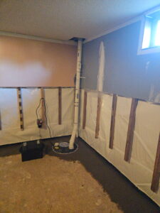 Basement corner with exposed walls and sump pump system. Pipes run vertically to the ceiling, with an electrical setup nearby. A small window provides light.