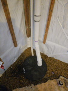 A basement corner with a sump pump system, featuring plastic pipes emerging from a circular black cover. The floor is lined with brown gravel, walls have moisture-proof sheeting.