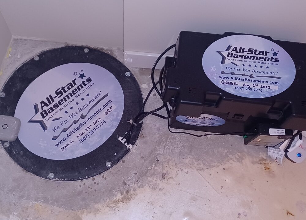 A sump pump with a black cover labeled "All-Star Basements" is installed in a basement floor. A battery backup system sits beside it on the concrete.