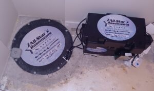 A sump pump with a black cover labeled "All-Star Basements" is installed in a basement floor. A battery backup system sits beside it on the concrete.
